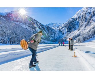 Man playing ice stock sport on a frozen lake, surrounded by snowy mountains and dense winter forest | © Rene Eduard Perhab