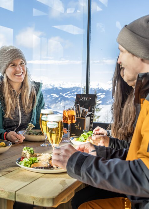 Four people in ski gear dining at a wooden table with mountain view in the restaurant | © Mirja Geh