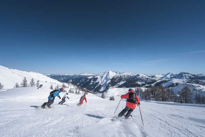 Three skiers and one snowboarder ride downhill together on a slope with snowy alpine peaks in the background.