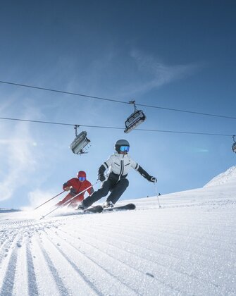 Two skiers ride side by side on groomed slope, above them a chairlift and bright sun shining through a clear sky.