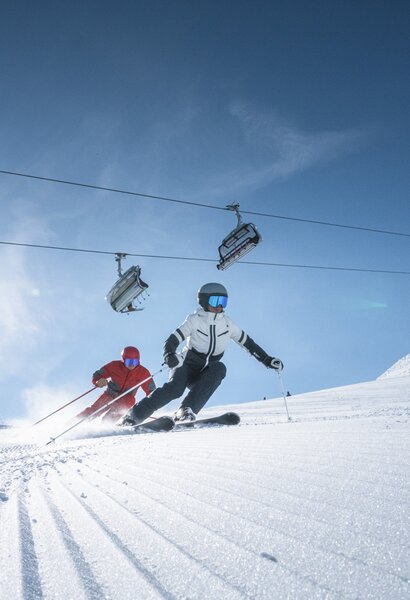 Two skiers ride side by side on groomed slope, above them a chairlift and bright sun shining through a clear sky.