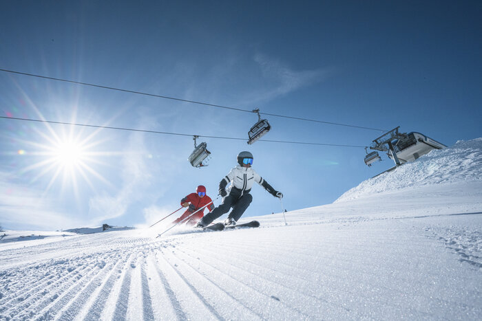 Two skiers ride side by side on groomed slope, above them a chairlift and bright sun shining through a clear sky.