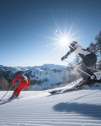 Two skiers carve down a freshly groomed slope, sunbeams shining through trees and above the mountain panorama.