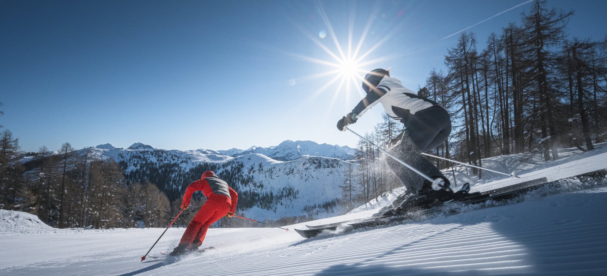 Zwei Skifahrer carven über frisch präparierte Piste, Sonnenstrahlen leuchten durch Bäume und über Berge.