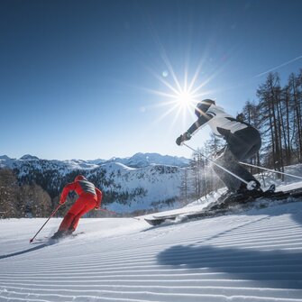 Two skiers carve down a freshly groomed slope, sunbeams shining through trees and above the mountain panorama.