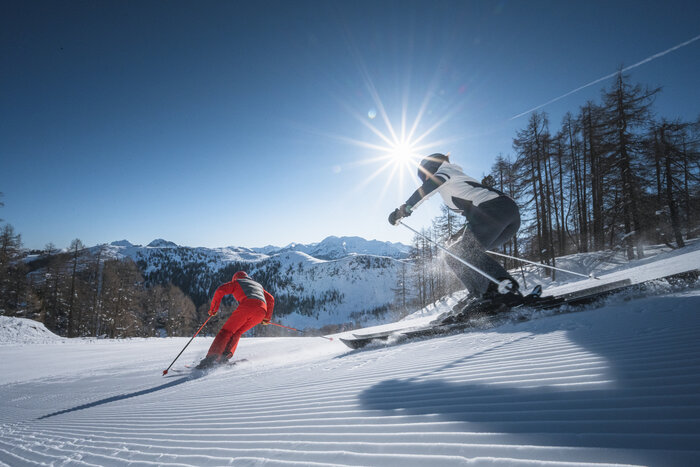 Zwei Skifahrer carven über frisch präparierte Piste, Sonnenstrahlen leuchten durch Bäume und über Berge.