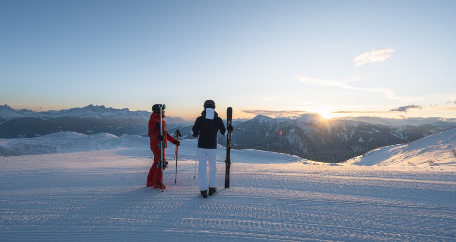 Two people in ski equipment stand on a piste and watch the sunrise