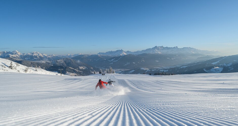 Two people ski down a freshly groomed slope and swirl up the snow