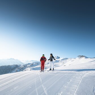 Two skiers stand side by side on freshly groomed slope with sunshine, chairlift and Alpine view