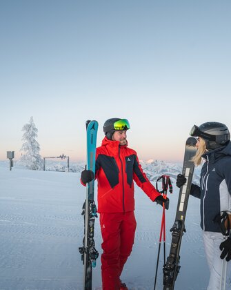 A man and a woman in ski gear stand with skis on a groomed slope in front of mountain scenery