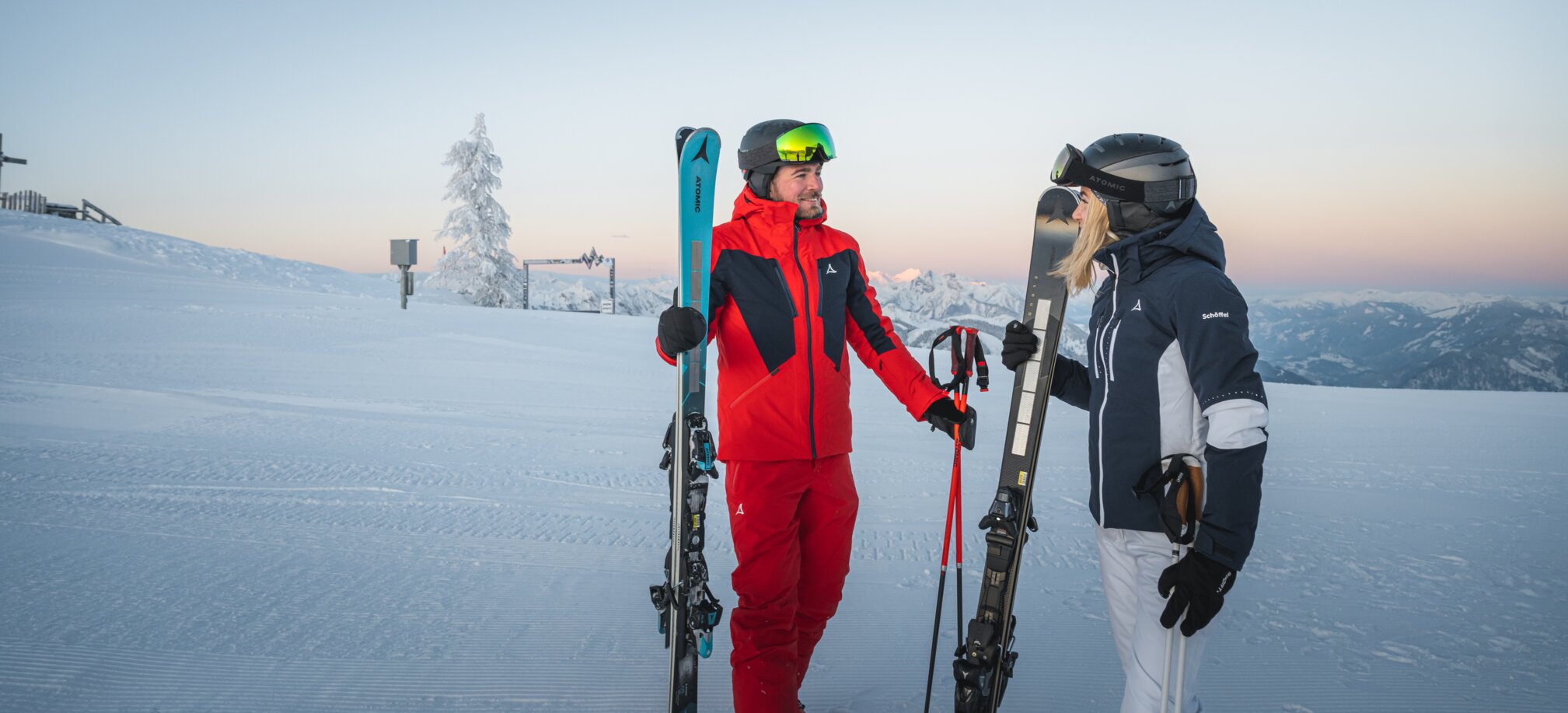A man and a woman in ski gear stand with skis on a groomed slope in front of mountain scenery