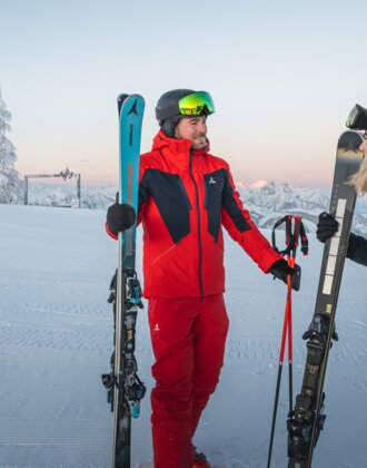 A man and a woman in ski gear stand with skis on a groomed slope in front of mountain scenery