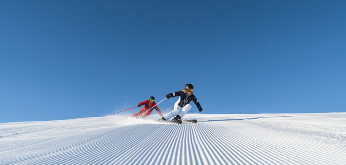 Two skiers skiing in low position on corduroy piste under cloudless blue sky
