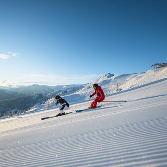 Two skiers skiing in parallel on a freshly groomed slope with sunshine and Alpine panorama in the background