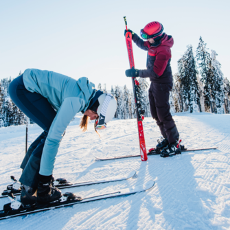 A skier bends down to her speedy skis by her ski boots and the skier is just looking at the unbuckled ski while he is still standing on the piste with the other one