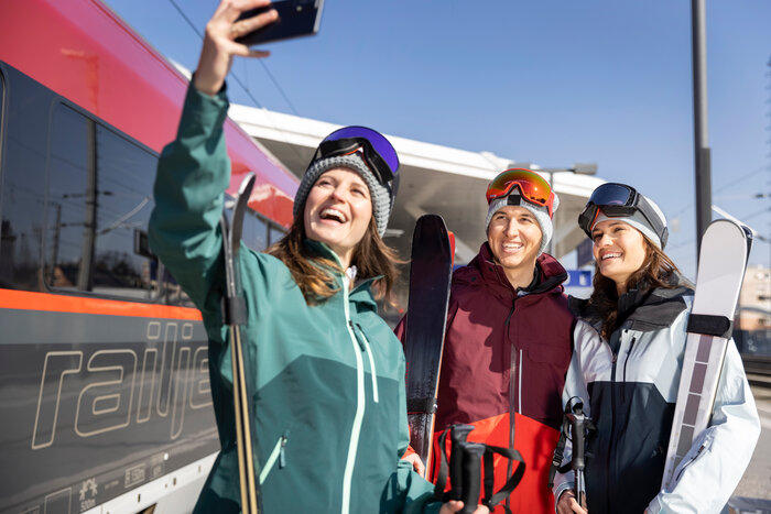 Three people in ski equipment stand on a platform next to a train and take a selfie | © ÖBB | Harald Eisenberger
